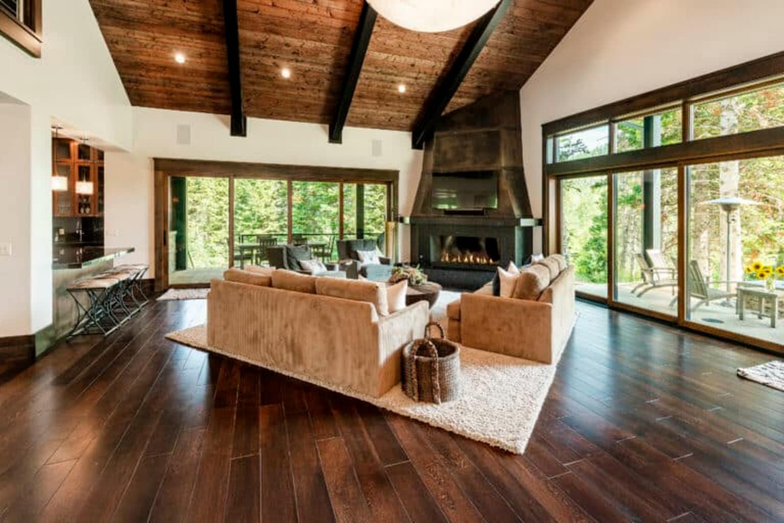 Spacious living room with vaulted wooden ceiling, large windows, beige sofas, fireplace, expertly refinished dark wood flooring by our Hardwood Floor Services, and outdoor patio view.
