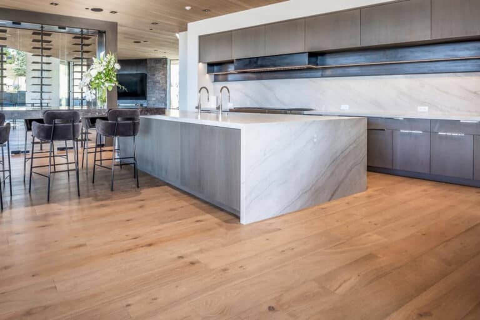 Modern kitchen with engineered wood flooring installation, a marble island with dual sinks, dark cabinetry, and bar stools; open shelving and large windows in the background—perfect for Park City UT living.