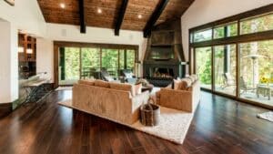 Modern living room with vaulted wood ceiling, large windows, dark hardwood floors, beige sectional sofa, fireplace, and a view of trees outside—see our 2024 Pricing Guide to compare the cost to replace carpet with hardwood.