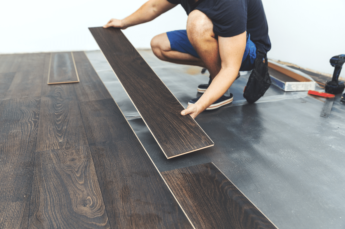 A person in shorts installs dark wood laminate flooring, aligning a plank on a prepared subfloor with tools nearby—showcasing expert Commercial Flooring Installation by Flooring Service Utah.