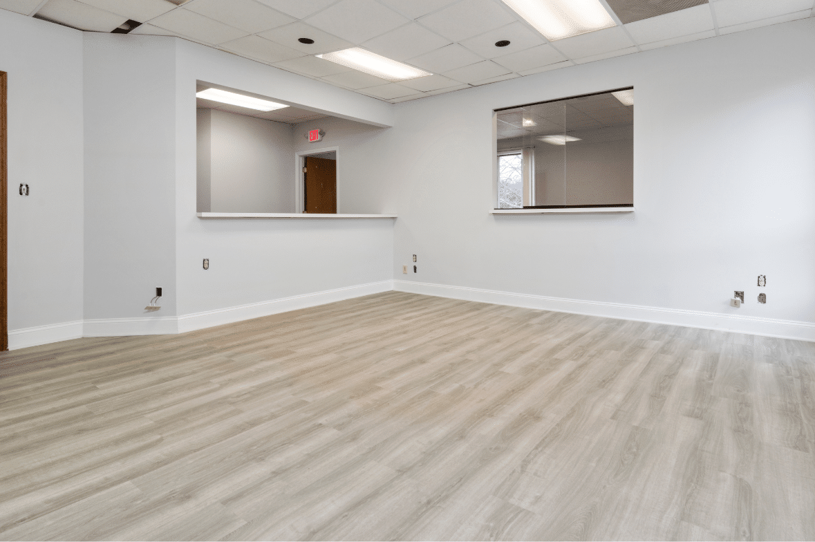 An empty office space with light wood flooring, white walls, a large interior window, drop ceiling tiles, and fluorescent lighting—perfect for businesses seeking quality commercial flooring installation in Utah.