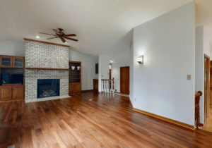 Living room with gorgeous hardwood floors—perfect for those considering hardwood floor refinishing—a white brick fireplace, built-in wooden shelves, vaulted ceiling with fan, and a wall sconce.