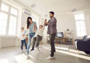 Two adults and two children play together in a bright living room with gleaming floors; the adults swing one child by the arms while the other smiles nearby, showing off the results of recent hardwood floor refinishing.