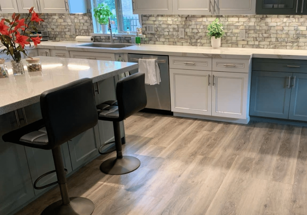 Modern kitchen with gray cabinets, a marble backsplash, and a large island with two black chairs. The light wood flooring adds warmth—perfect if you're considering hardwood floor replacement or refinishing to elevate your space. Plants thrive near the window.