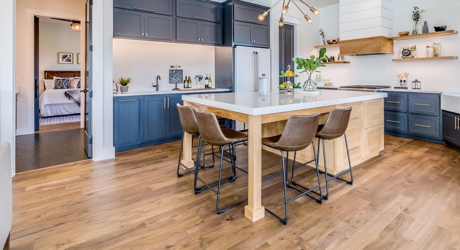Modern kitchen with blue cabinets, wooden island with four stools, pendant lighting, and expertly refinished hardwood floors. A bedroom is visible through an open doorway, showcasing the quality provided by professional Floor Refinishing Services.