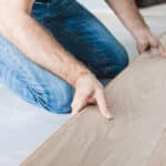 A person installs light wood laminate flooring, aligning the planks by hand. The logo "Accent Flooring" is visible in the bottom right corner.