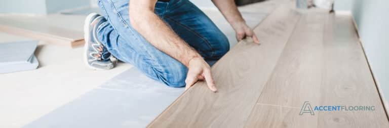 A person installs light wood laminate flooring, aligning the planks by hand. The logo "Accent Flooring" is visible in the bottom right corner.