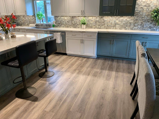 Modern kitchen with trending flooring in light wood, blue and white cabinets, stone backsplash, island with black stools, and a dining area featuring upholstered chairs.