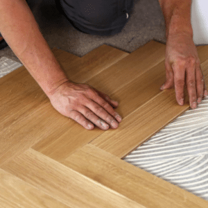 A person installs wooden floor panels, aligning them over adhesive on a subfloor—the perfect step before you refinish hardwood floors or decide when is the best time for hardwood floor refinishing.